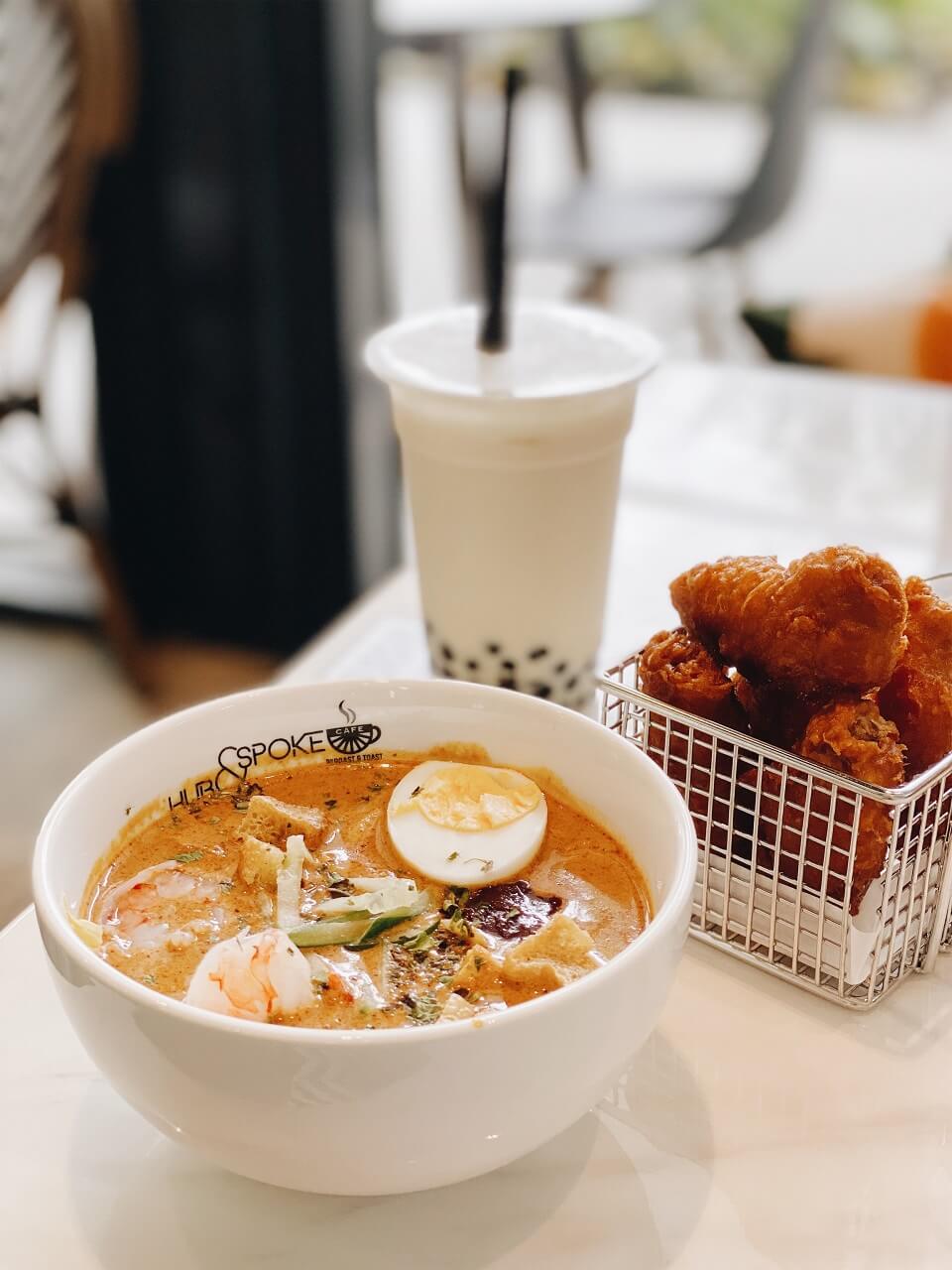 A bowl of laksa with a side and bubble tea in the background.