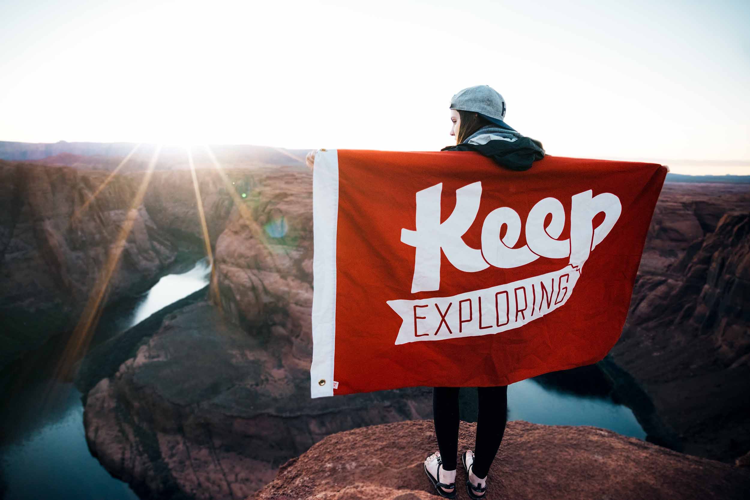 A woman standing on top of a mountain with a cape with the words 'Keep Exploring'