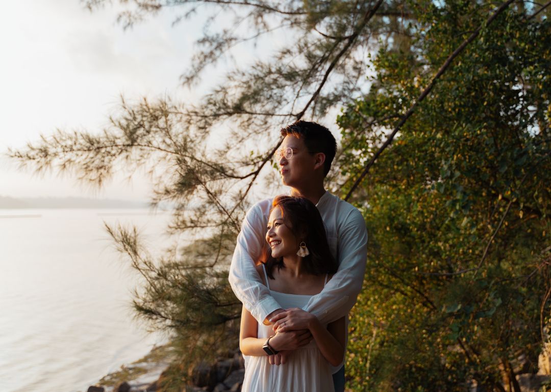 A groom holds his bride from the back at dawn as they gaze at the waters with soft trees in the background.