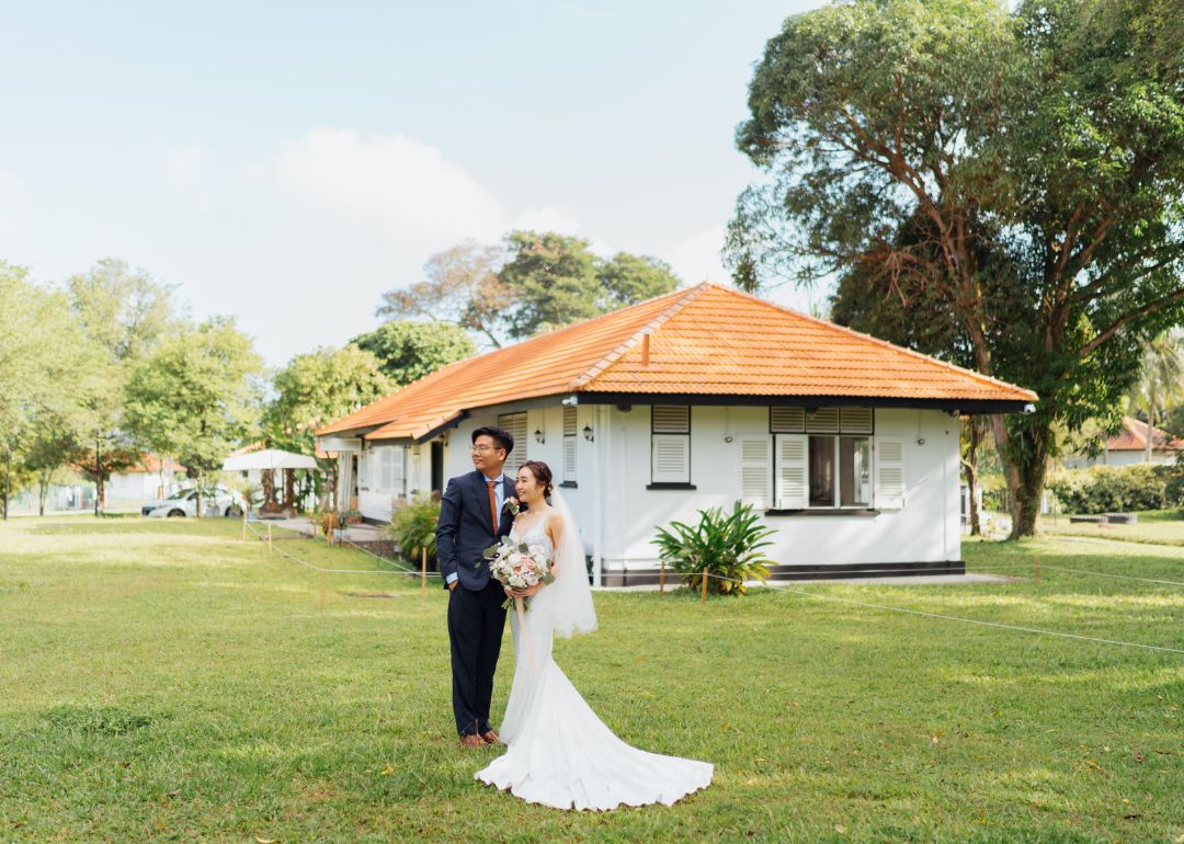 A wedding couple on a lawn standing in front of a colonial bungalow in an outdoor location.