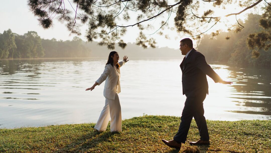 A wedding couple standing apart looks at each other with their arms outstretched as they  bask in the scenery in front of the reservoir at dawn.