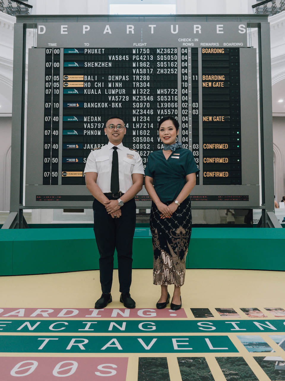 two uniformed staff – a man and a woman – stand before the solari board at the now boarding travel exhibition